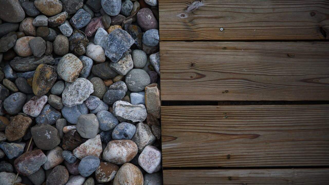 Collection of smooth river rocks in various colors beside weathered wooden planks, showing materials for hardscaping landscaping projects