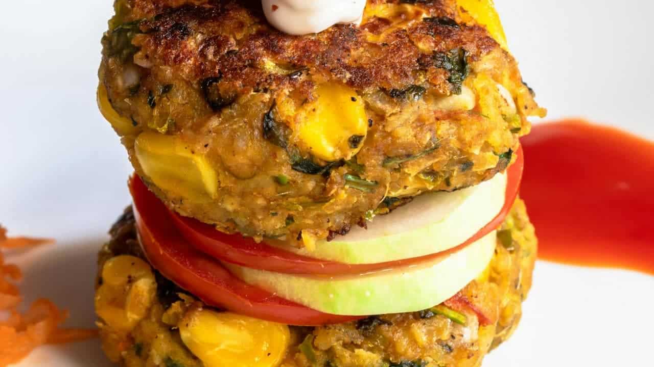 Stacked corn fritters made with visible corn kernels, herbs, and spices, layered with fresh tomato and green apple slices, topped with a dollop of sauce, served with a side of red dipping sauce