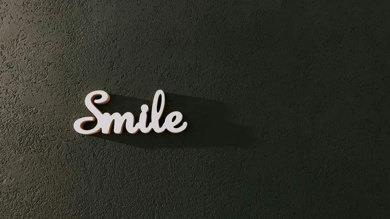 Word "Smile" in cursive white lettering mounted on a dark textured wall, casting a soft shadow to the right, minimalistic decor with contrast between light and surface texture
