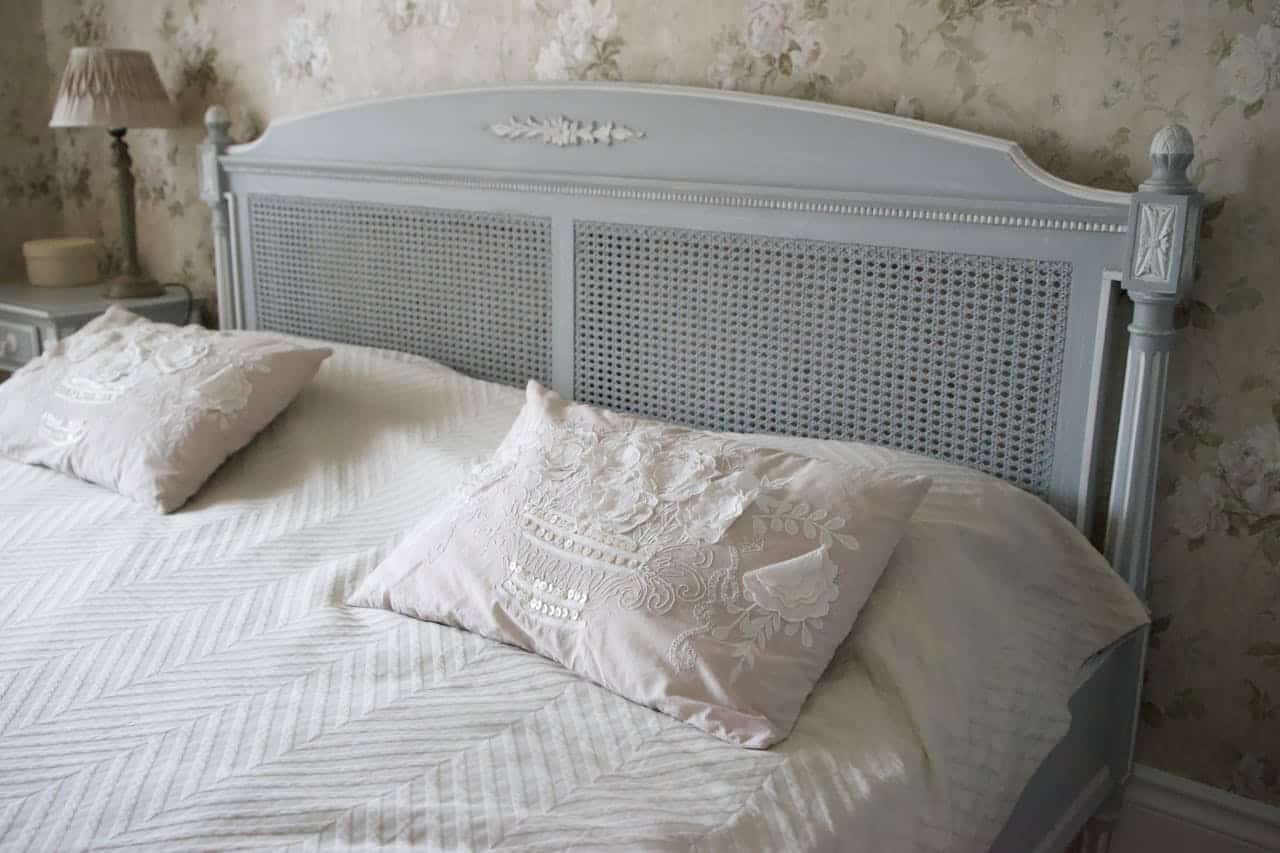 Light blue wooden bed with cane headboard, white bedding, embroidered vintage linens pillows, against floral wallpaper in vintage-style bedroom