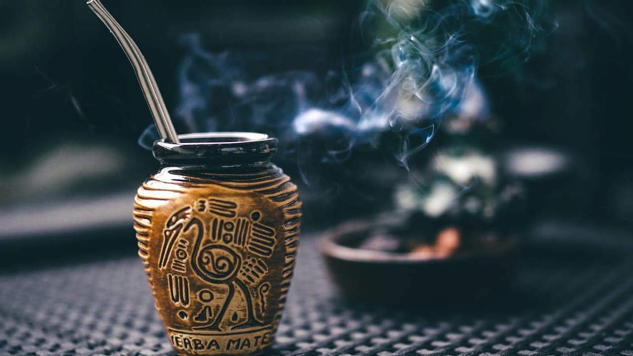 Traditional carved wooden smoke infuser with metal straw and rising steam, sitting on textured surface with blurred background