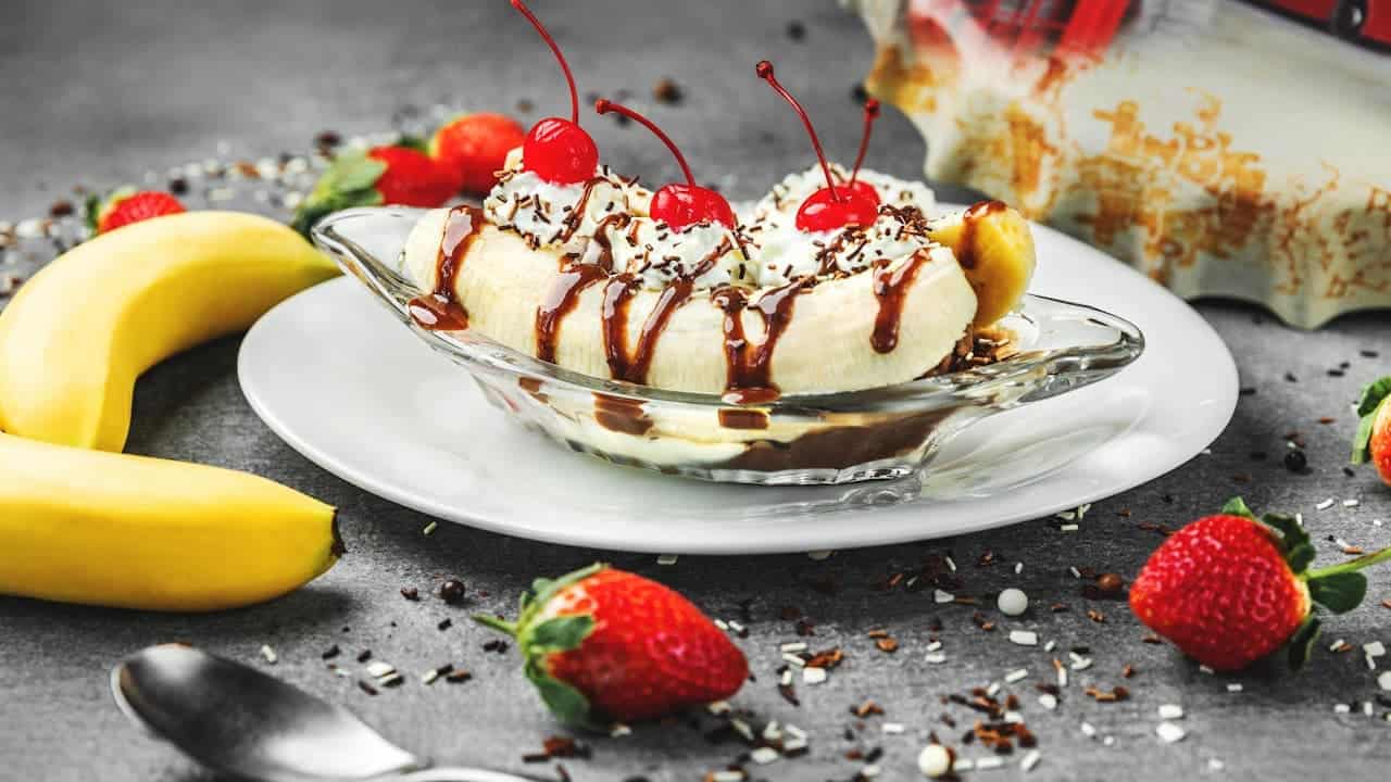 Banana split dessert with whipped cream, chocolate syrup, and maraschino cherries, surrounded by fresh strawberries and bananas