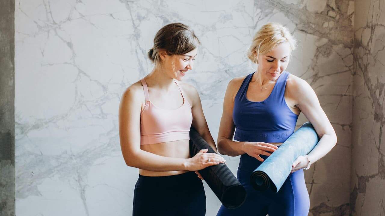 Two women in athletic wear holding yoga mats, standing in front of a marble wall, smiling and looking at each other