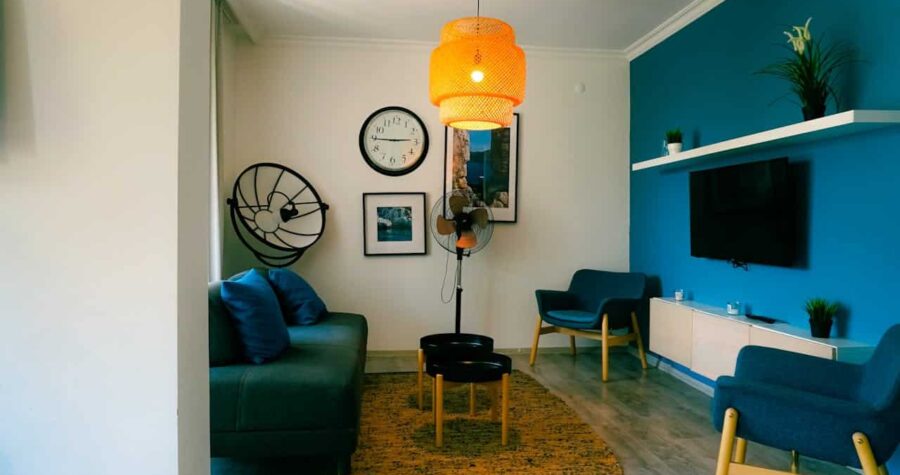 Cozy living room with teal accent wall, orange pendant light, blue furniture, standing fan, wall clock, and framed artwork