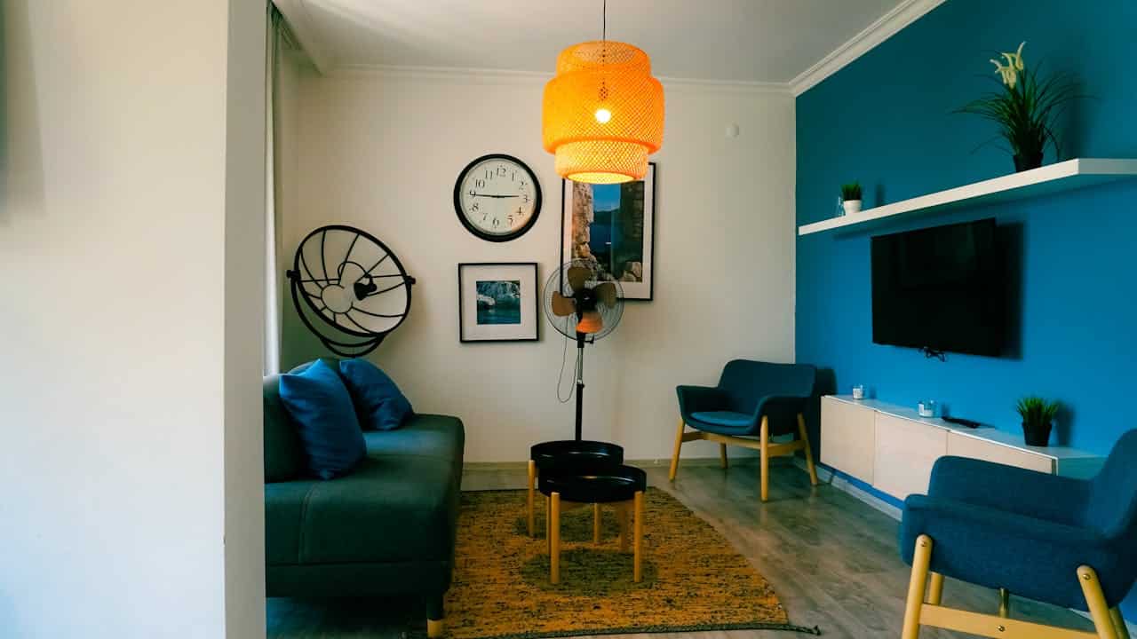 Cozy living room with teal accent wall, orange pendant light, blue furniture, standing fan, wall clock, and framed artwork