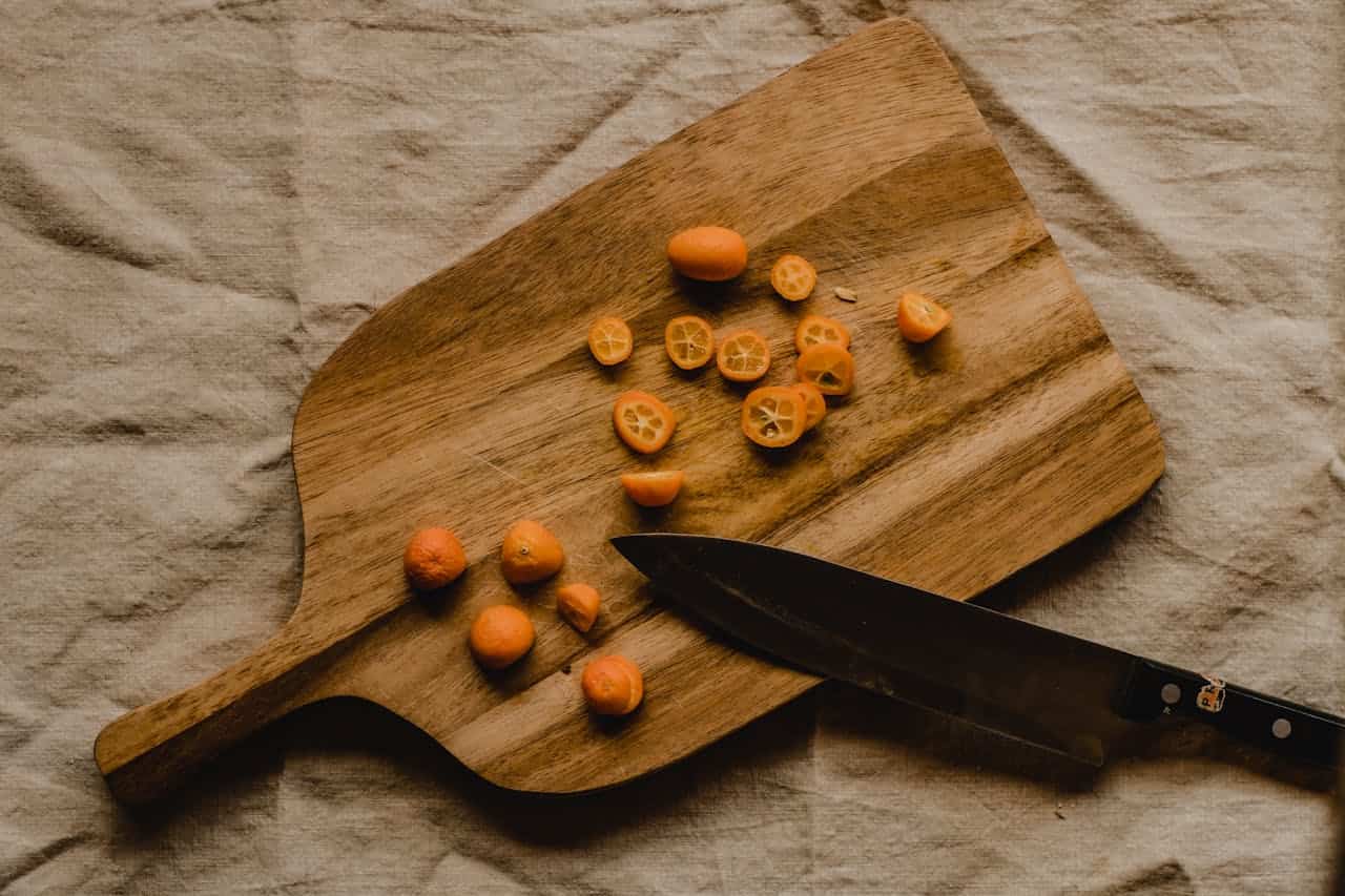 Wooden cutting board with sliced kumquats and a chef's knife on beige linen cloth background
