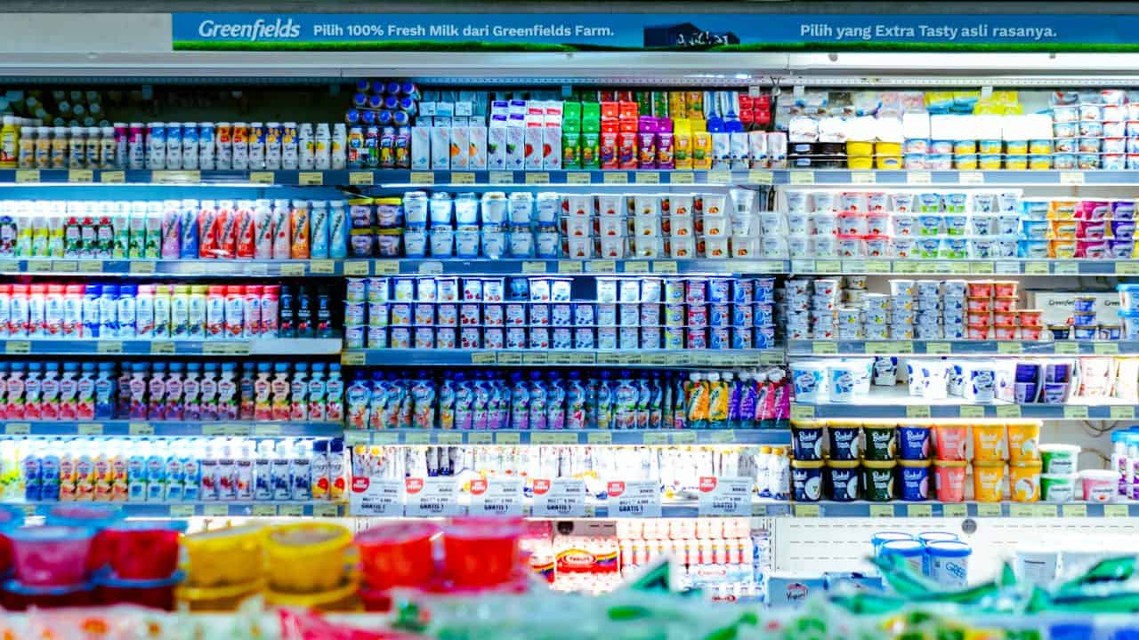 Supermarket refrigerated section with colorful dairy products, featuring Greenfields milk banner in Indonesian language above densely stocked shelves