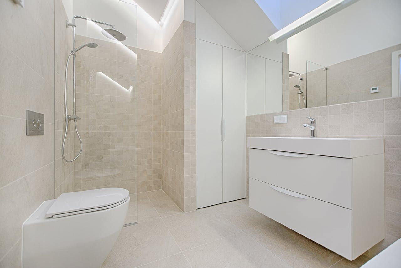 Modern bathroom with beige tiles, walk-in shower with rainfall and handheld heads, wall-mounted toilet, large vanity with drawers, wide mirror, and concealed storage cabinet under sloped ceiling