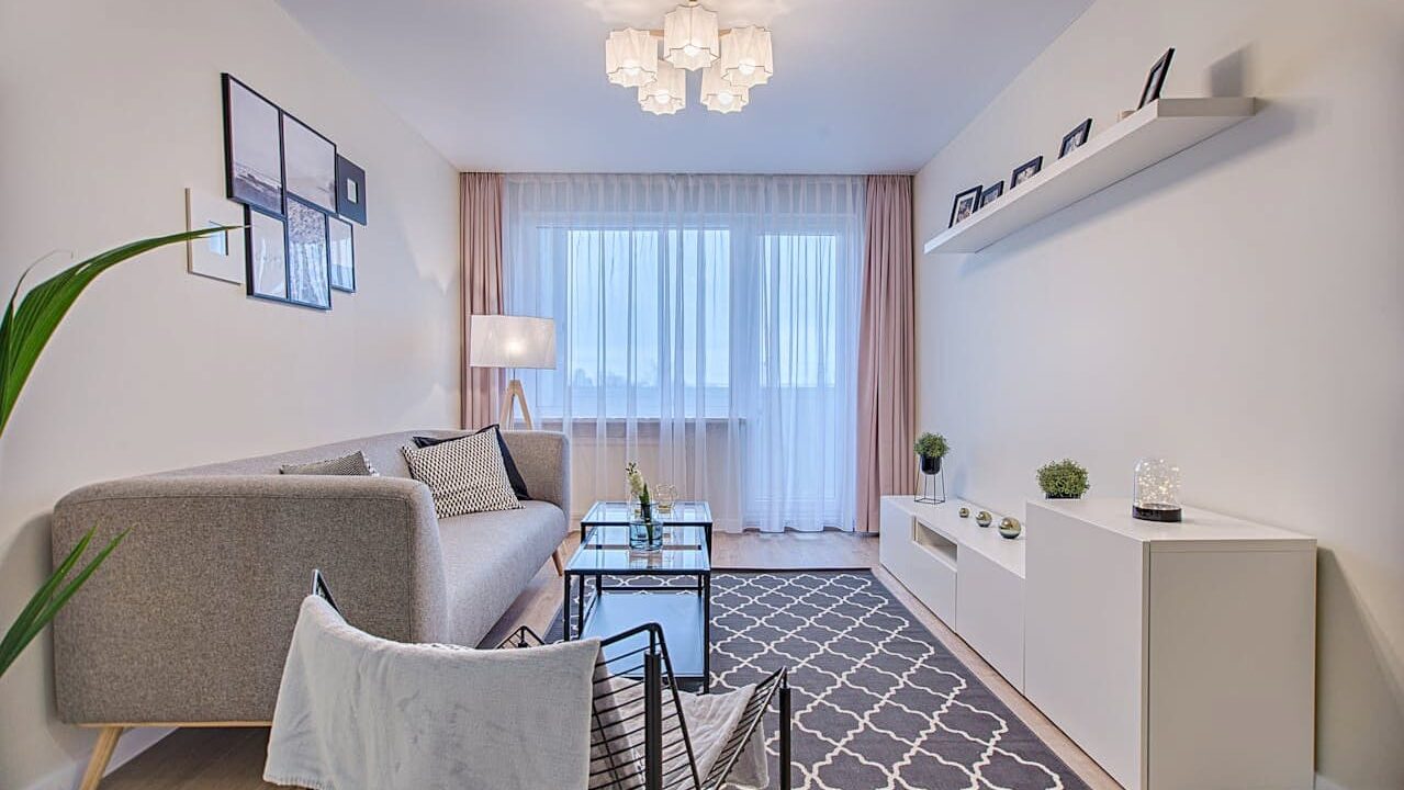 Compact modern living room with gray sofa, white media console, glass coffee table, white walls, patterned rug, and pink curtains