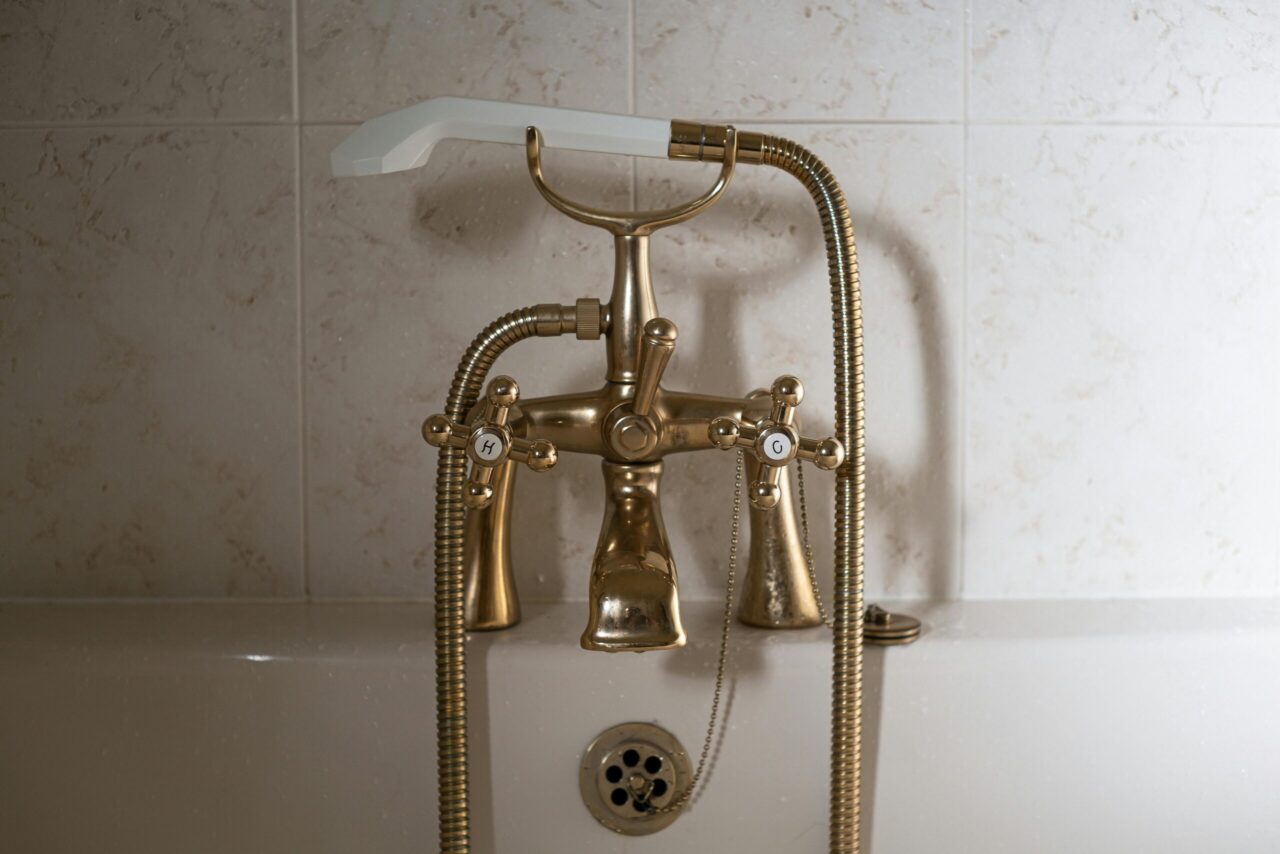 Vintage-style bathtub faucet, brass finish, labeled hot and cold handles, flexible shower hose attached, white wall tiles in the background, metal drain plug with chain, classic bathroom fixture design