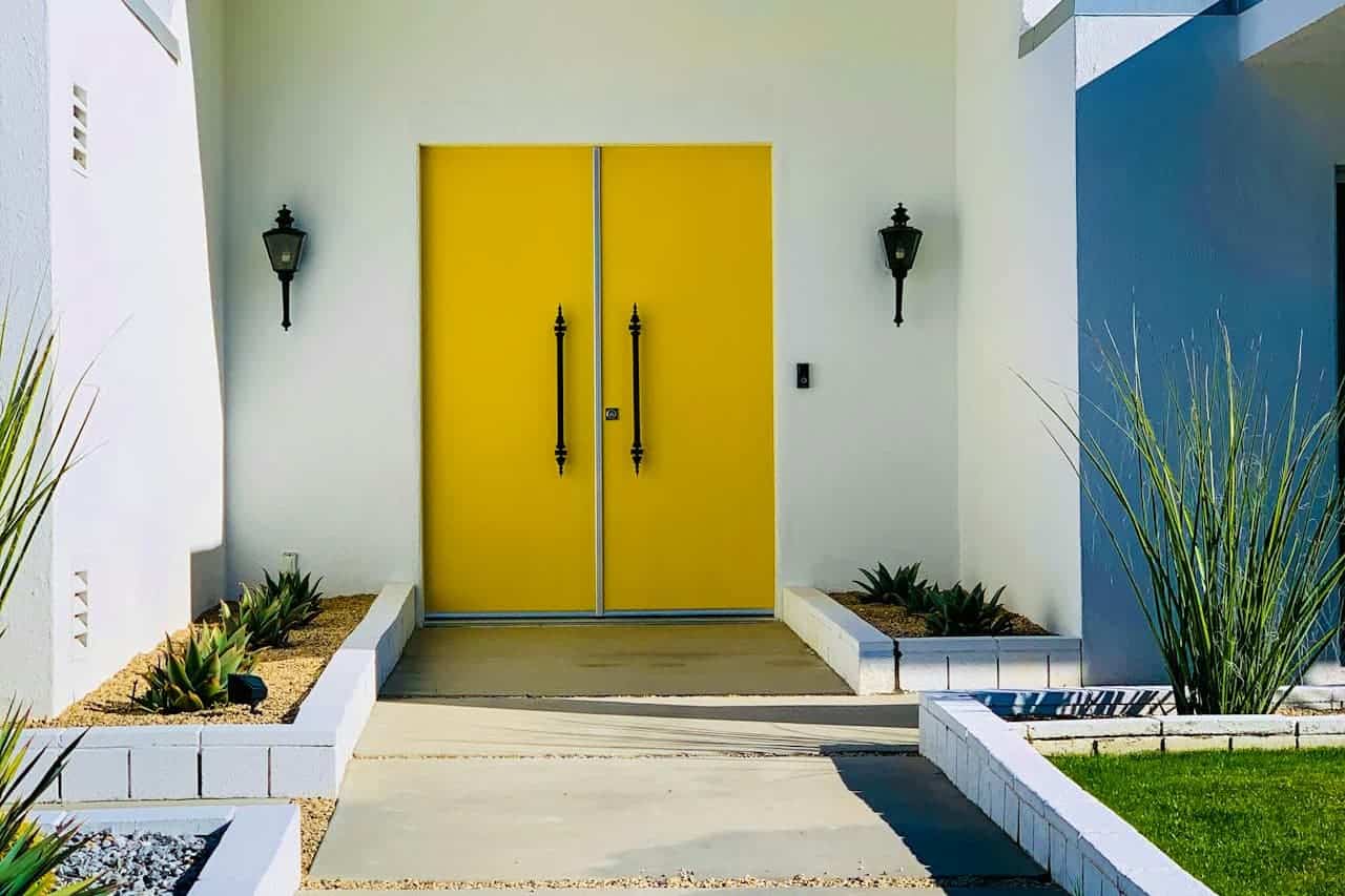Modern home entrance featuring bright yellow double doors, white walls, black sconces, and drought-resistant landscaping with succulents