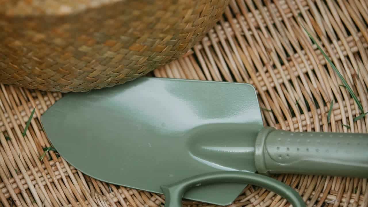 Close-up of a green gardening trowel with a textured handle resting on a woven wicker surface, partially covered by a woven straw hat