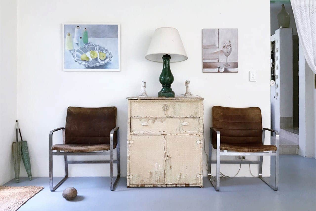 Vintage interior with two brown leather chairs flanking distressed cream cabinet, green lamp, wall art, and umbrella stand