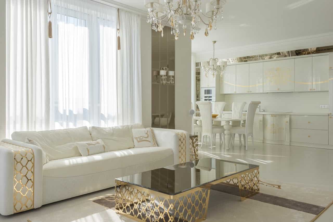 Luxurious white and gold open-concept living room with crystal chandelier, white sofa, glass coffee table, and modern kitchen