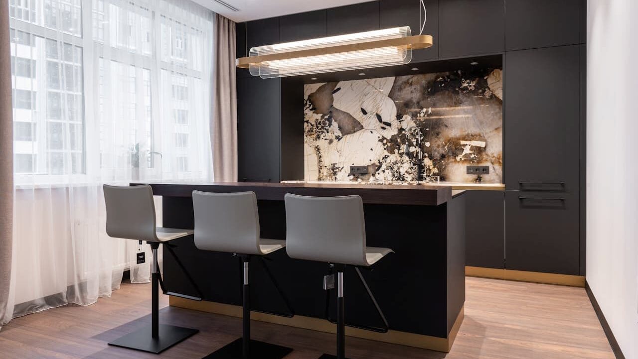 Kitchen with matte black chalk paint cabinetry, marble-pattern backsplash, wooden countertop, three white barstools, glass pendant light, and sheer window curtains