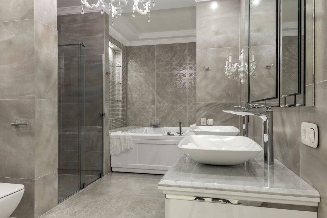 Luxurious gray bathroom with white vessel sink, marble countertop, glass shower enclosure, soaking tub, and crystal chandelier