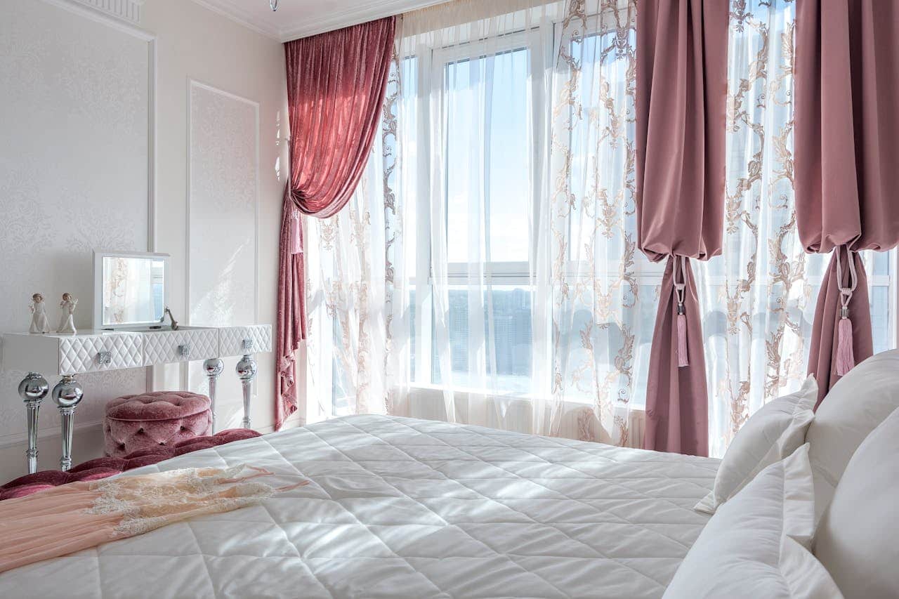 Elegant bedroom with white quilted bed, rose pink curtains, sheer embroidered drapes, vanity table, and bright windows