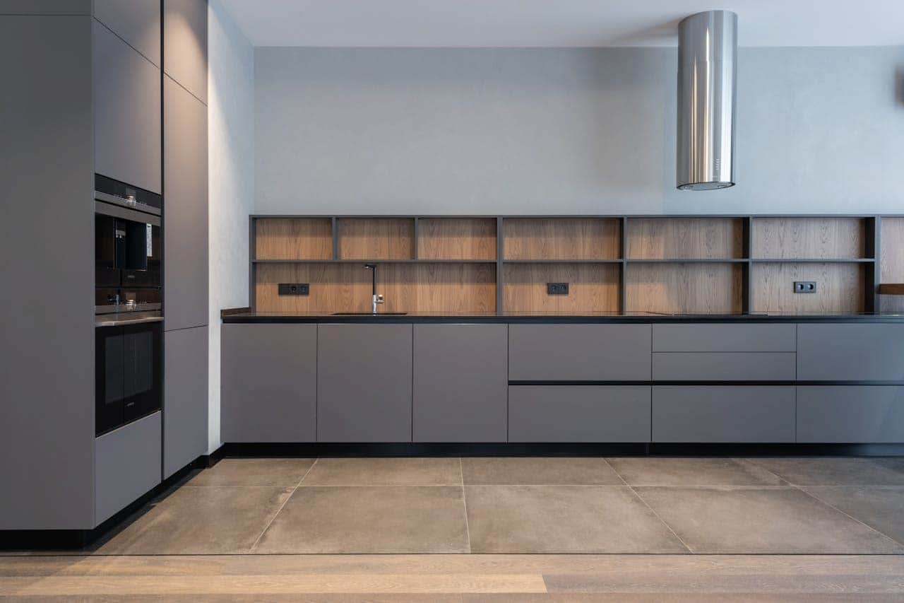 Minimalist modern kitchen with gray cabinetry, wooden backsplash shelving, stainless steel hood, built-in appliances, and travertine tile flooring