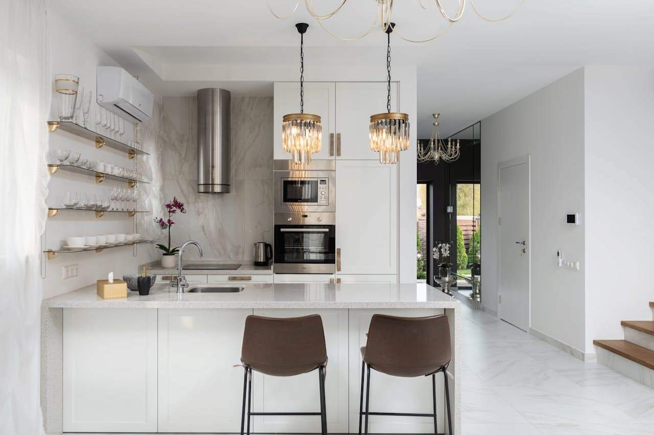 Modern white kitchen with marble backsplash, copper accents in lighting, brown barstools, open shelving, and stainless steel appliances