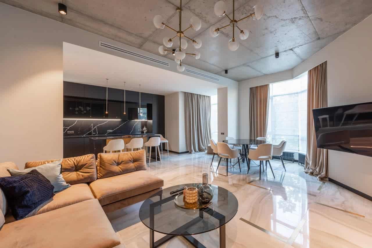 Modern open-concept apartment with Bauhaus and loft styles, with beige sectional sofa, glass coffee table, dining area, black kitchen, and concrete ceiling with pendant lights