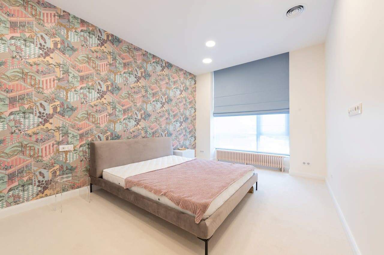 Minimalist bedroom with gray upholstered bed, pink throw blanket, and feature wall with colorful architectural pattern wallpaper