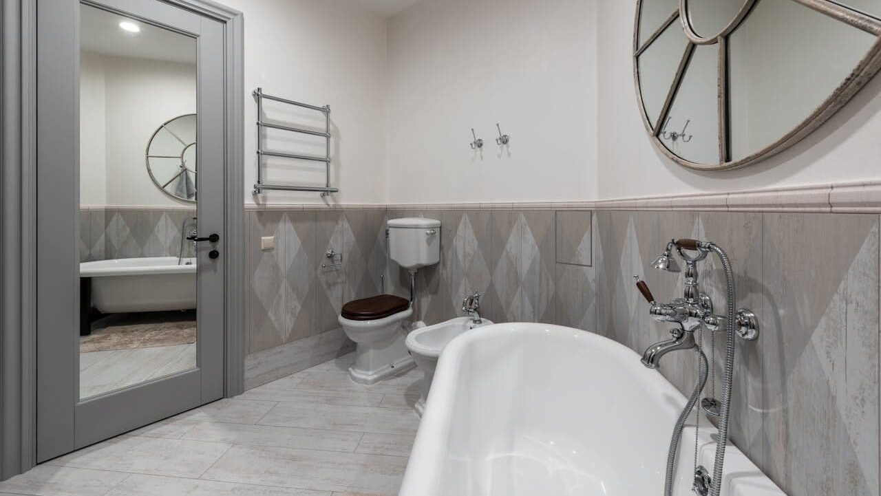Elegant bathroom featuring white freestanding bathtub, toilet with wooden seat, diamond-patterned wall tiles, towel warmer, and decorative mirror