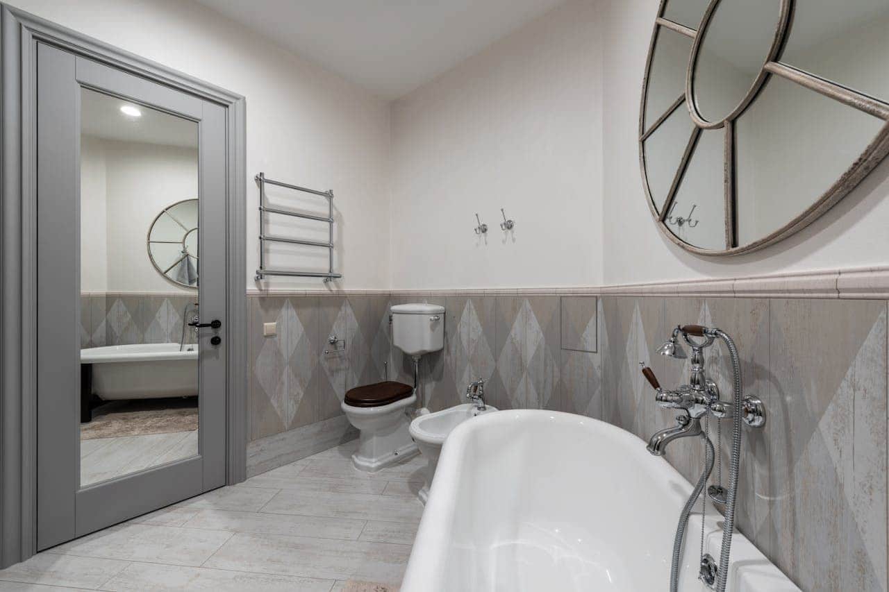 Elegant bathroom featuring white freestanding bathtub, toilet with wooden seat, diamond-patterned wall tiles, towel warmer, and decorative mirror