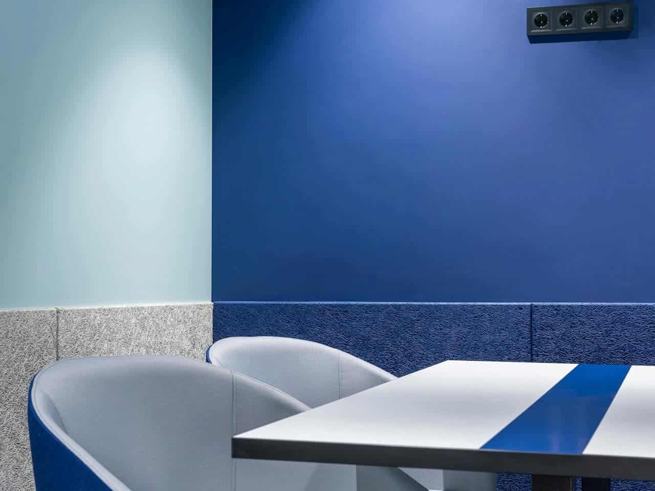 Modern interior with a blue and green wall, textured grey base, a minimalist table with a blue stripe, two comfortable grey chairs, a power outlet above the table, clean and contemporary design, suitable for a relaxed setting