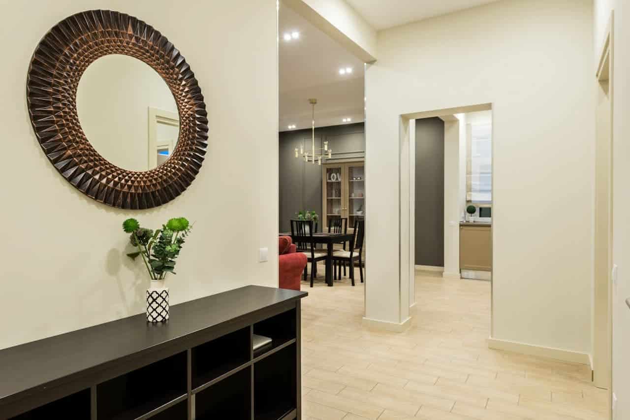 Entryway with dark console table, decorative round mirror, patterned vase with greenery, beige tiled floor, and view into dining area