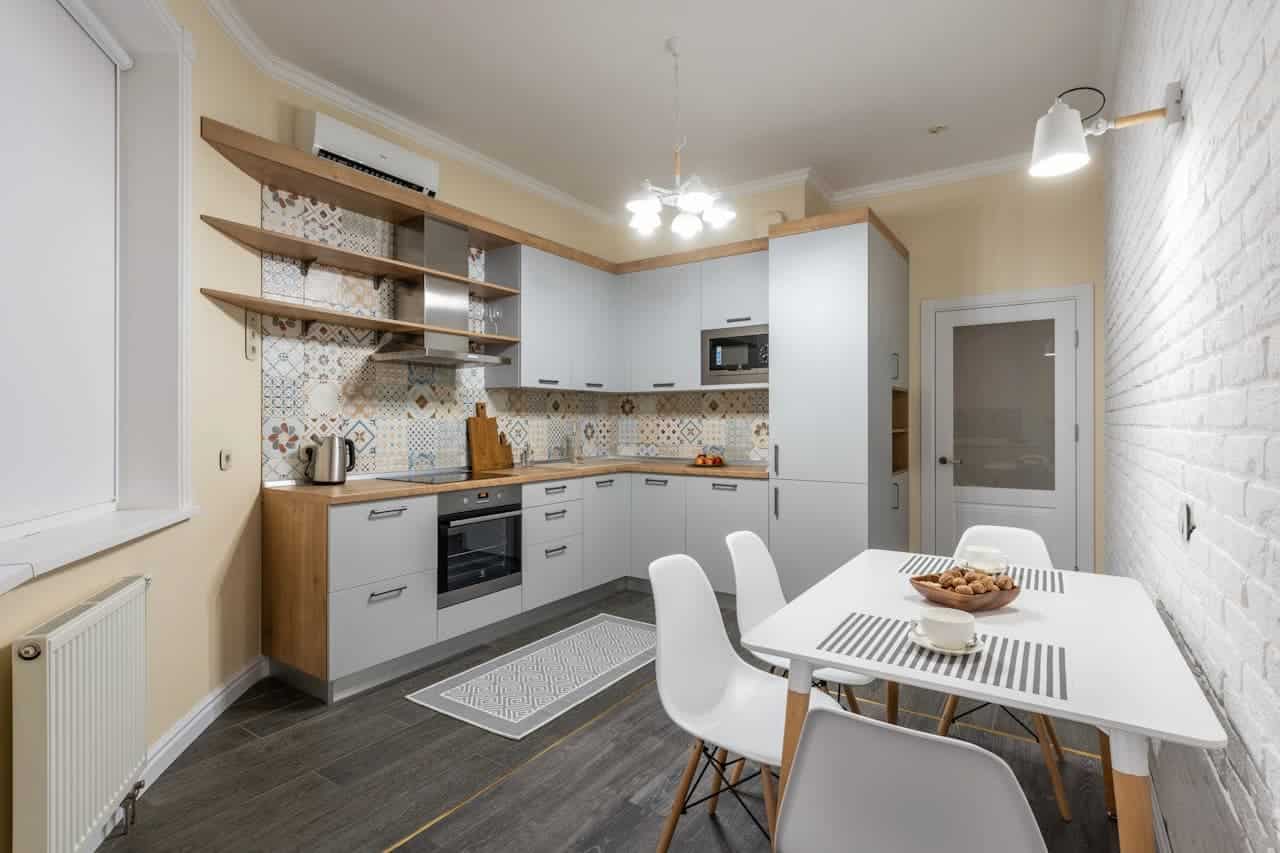 Modern L-shaped kitchen with white cabinets, patterned backsplash, dining table with chairs, white brick wall, and warm ambient lighting