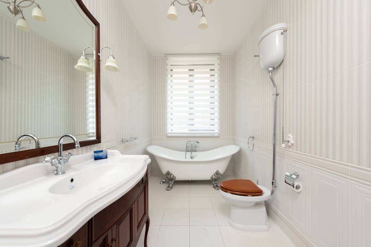 Elegant white bathroom with clawfoot tub, wooden vanity, toilet with wooden seat, fluted ceramic tiles, and window with blinds
