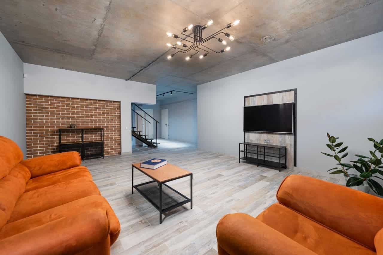 Modern industrial living room with orange sofas, concrete ceiling, exposed brick wall, wooden floors, and wall-mounted television