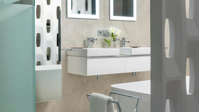 Modern bathroom with floating double sink vanity, illuminated mirrors, freestanding bathtub, frosted glass shower door, geometric white partition, and a metal towel rack with a white towel
