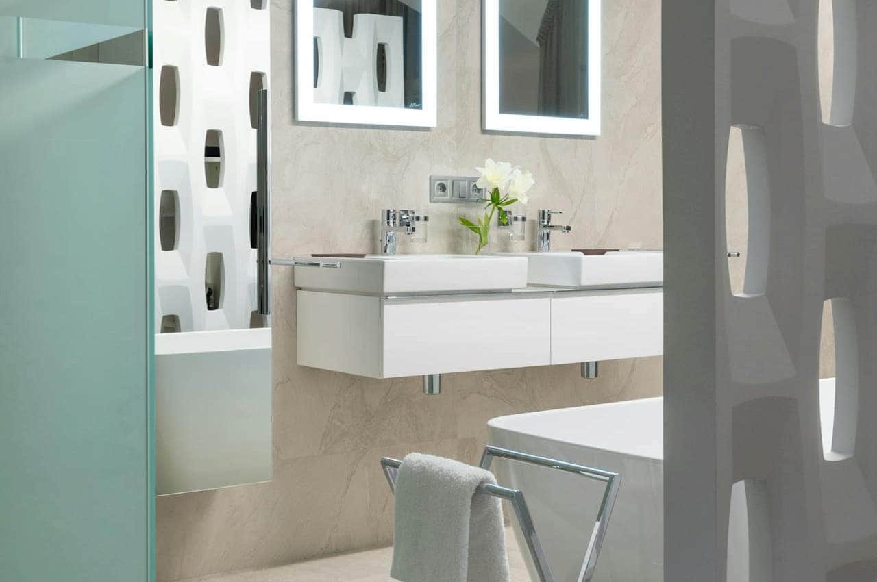 Modern bathroom with floating double sink vanity, illuminated mirrors, freestanding bathtub, frosted glass shower door, geometric white partition, and a metal towel rack with a white towel