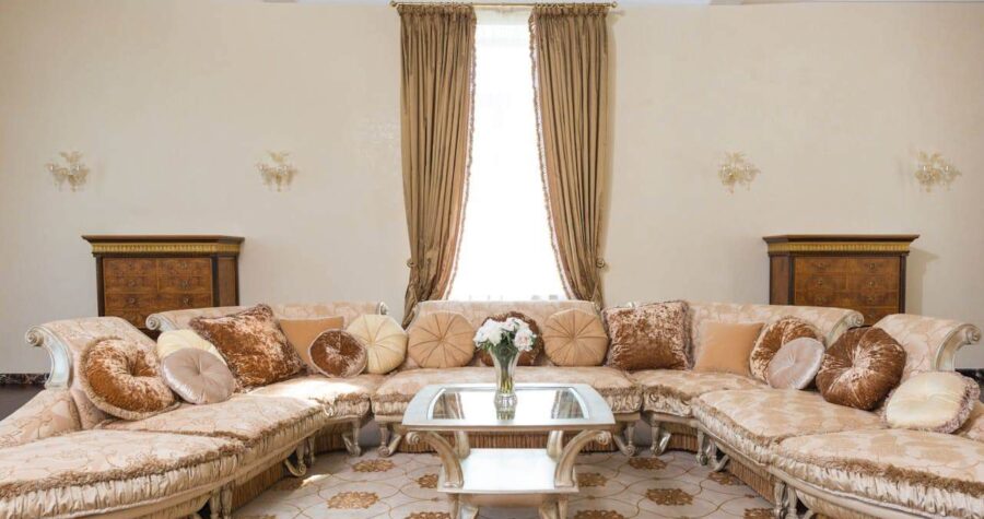 Luxurious cream-colored sectional sofa with decorative pillows in elegant living room featuring glass coffee table and beige curtains