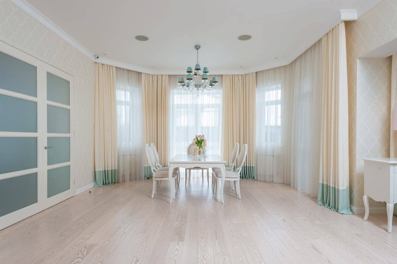 Elegant dining room with white table and chairs, cream and mint curtains, crystal chandelier, whitewashed floors, white ceiling and bay windows