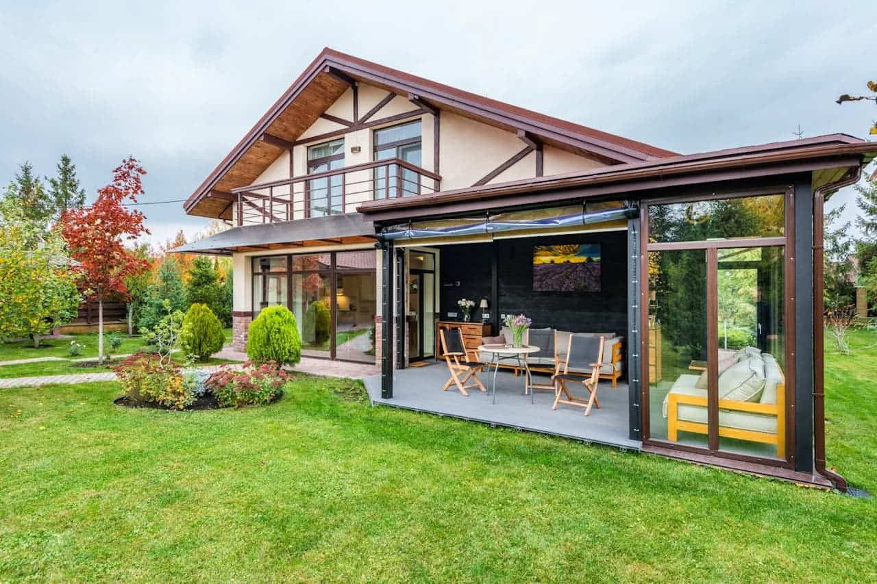 Modern farmhouse with Tudor-style elements featuring glass-enclosed patio, wooden furniture, manicured lawn, and colorful autumn landscaping