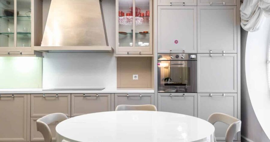 All-white kitchen with cabinetry, built-in appliances, stainless hood, glass display cabinet, and round white dining table with chairs