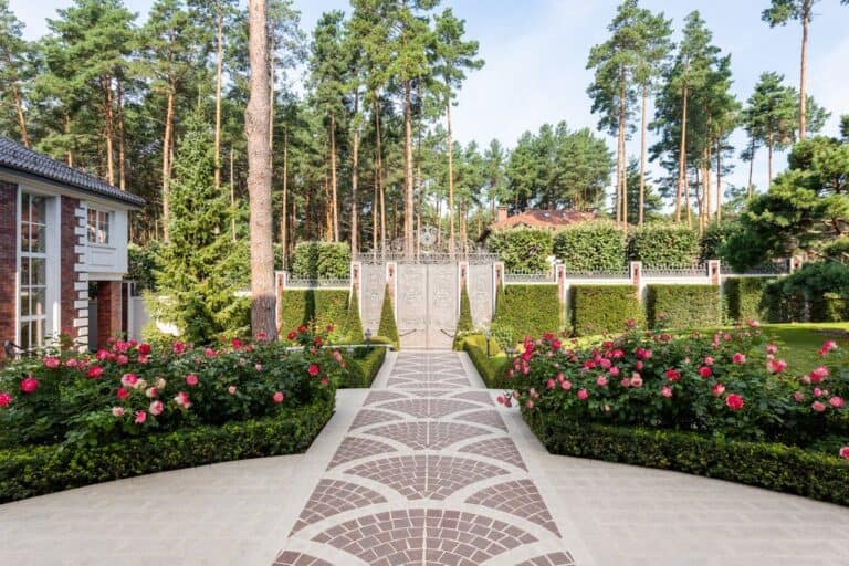 Elegant garden pathway with pink roses and trimmed low growing boxwood hedges leading to ornate gate, surrounded by tall pine trees