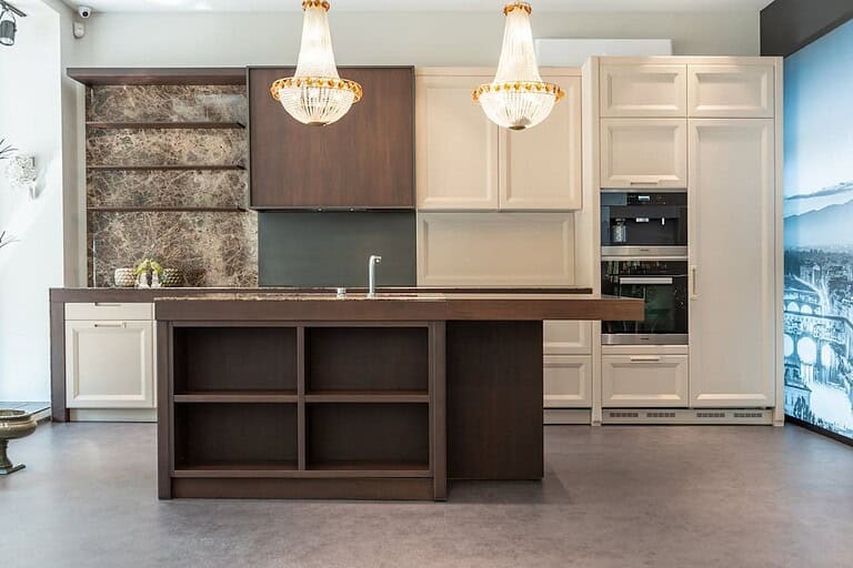 Modern kitchen with two-tone cabinetry, brown and cream, featuring crystal chandeliers, marble backsplash, island with shelving, and built-in appliances