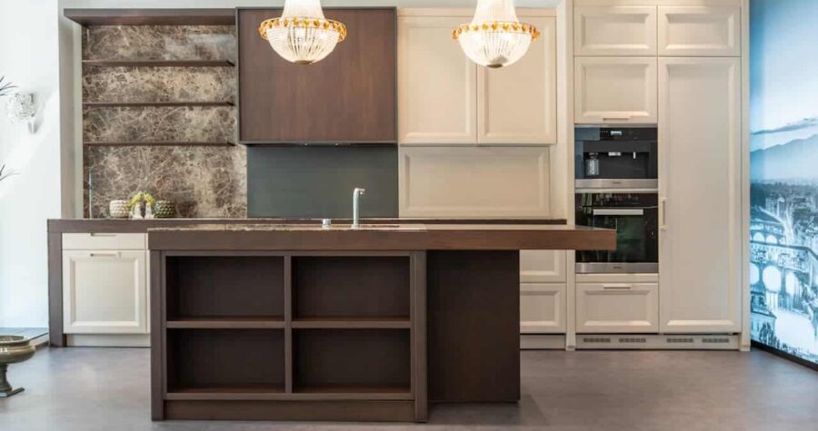 Modern kitchen with two-tone cabinetry, brown and cream, featuring crystal chandeliers, marble backsplash, island with shelving, and built-in appliances