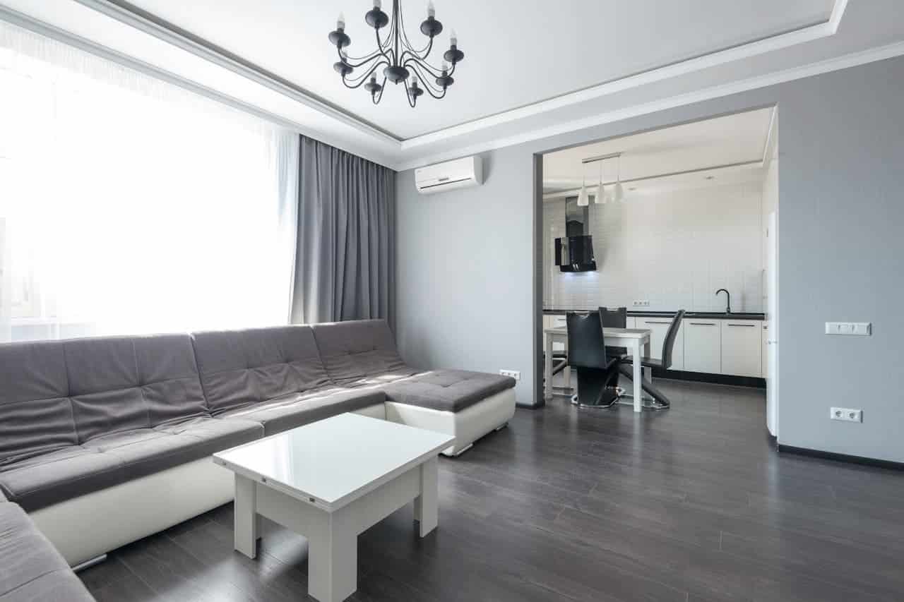 Modern gray and white apartment living room with sectional sofa, coffee table, black chandelier, and open kitchen view