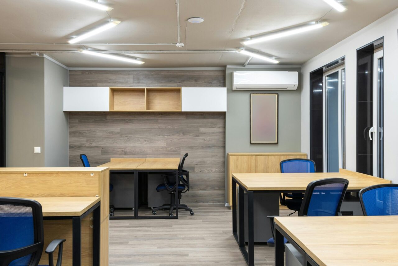 Wall-mounted air conditioner, installed above a framed picture, in a modern office with wooden desks, blue chairs, LED ceiling lights, large windows, and a clean layout