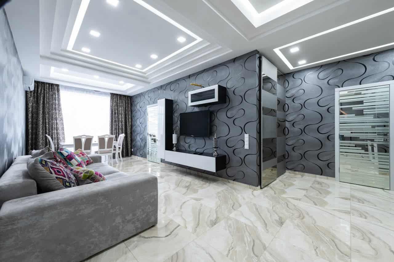 Modern living room with gray sectional sofa, textured black wallpaper walls, white entertainment center, marble floors and illuminated tray ceiling
