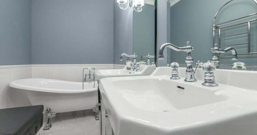 A luxurious bathroom with a white freestanding bathtub, a sleek white sink with chrome faucets, and a large mirror reflecting the space, accented by elegant chrome fixtures, a modern towel rack, and a minimalist black bench, all set against soft blue walls and glossy marble floors