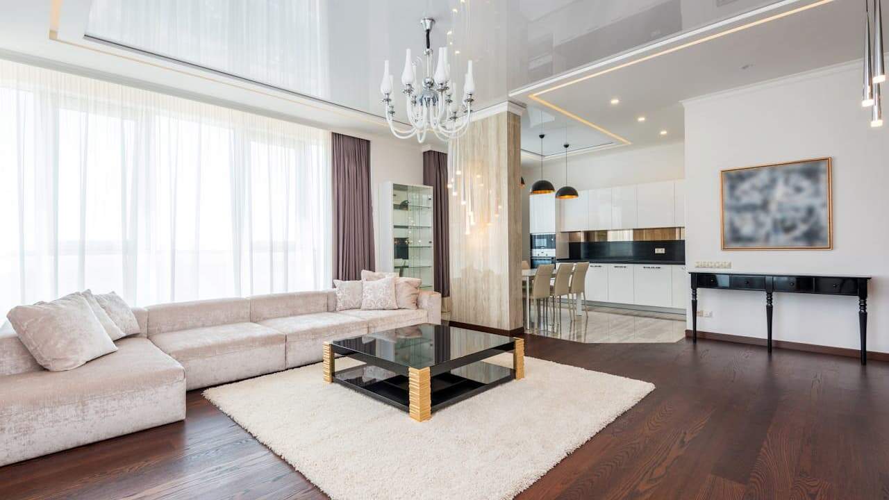 Open-concept layout living room with beige sectional sofa, glass coffee table, crystal chandelier, and open-plan kitchen with white cabinets