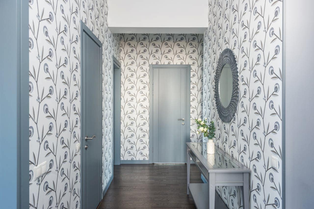 Elegant hallway with floral patterned wallpaper, light blue doors, dark wood floor, console table, and decorative oval mirror
