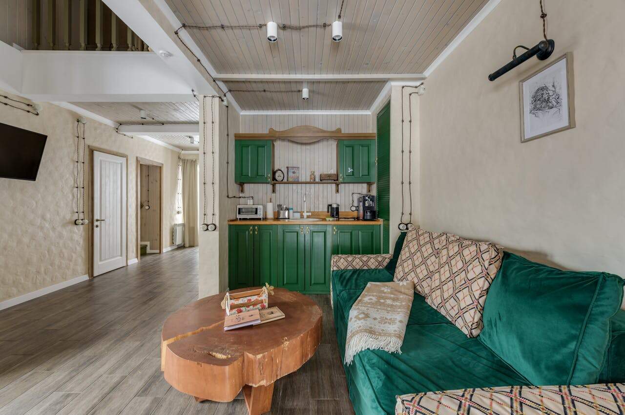 Cozy living space with rustic decor, green kitchen cabinets, wood countertop with appliances, green velvet sofa with patterned pillows and throw, round wooden coffee table with books, light wooden flooring, cream walls with visible wiring, ceiling lights