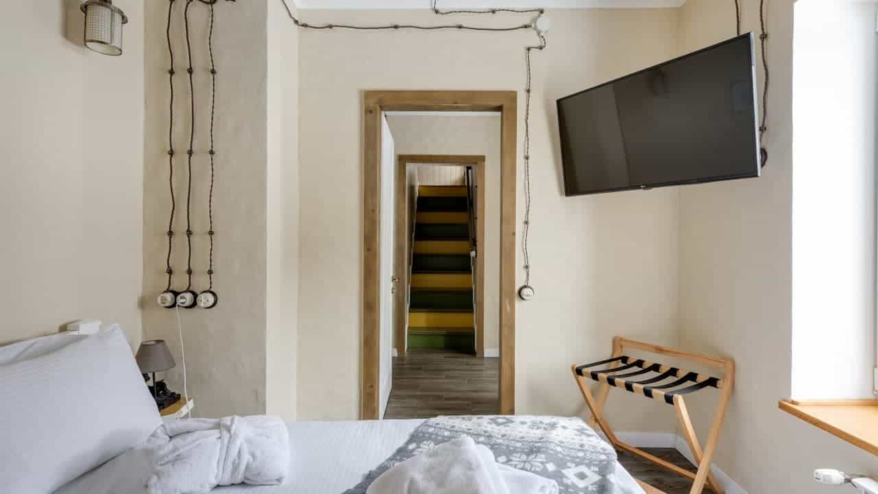 Modern hotel bedroom with white bedding, wall-mounted TV, pendant lighting, wooden luggage rack, visible wires and doorway leading to colorful staircase