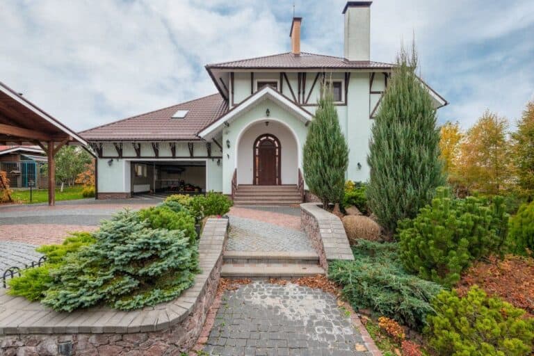 White Tudor-style house with arched entrance, landscaped pathway, evergreen shrubs, and cobblestone driveway under cloudy autumn sky